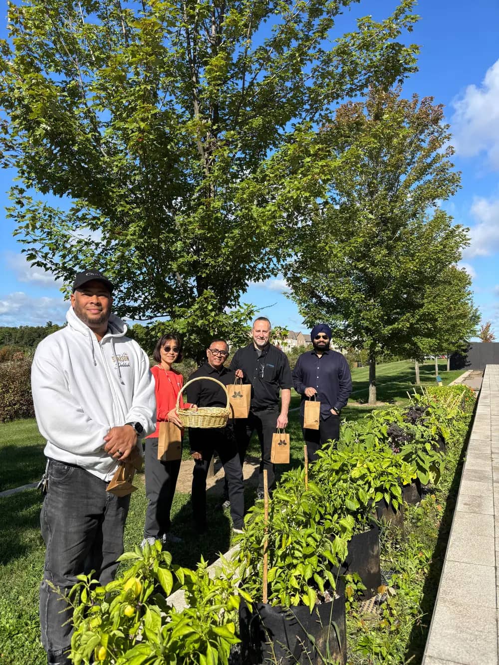 Five people stand behind planters holding gift bags and a basket on a sunny day