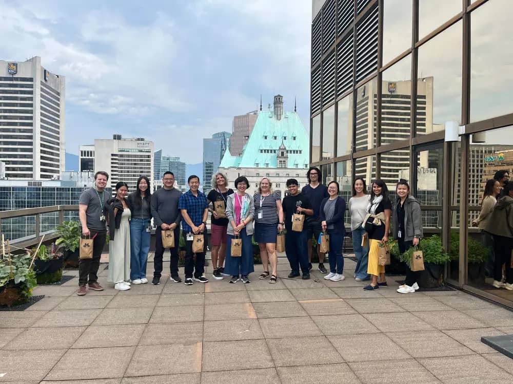 A group of people stand on a rooftop terrace holding paper bags with a city skyline behind them