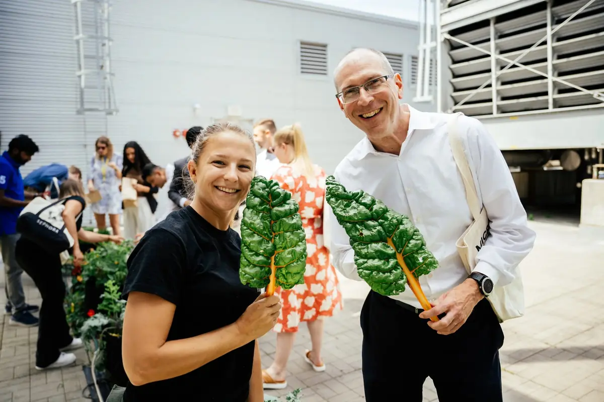 A farmer and a man in a white shirt each hold a chard leaf and smile
